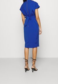 Blå klänning med korta ärmar, figursydd midja och knälång nederkant. Har en bakre knytning och dragkedjedetalj. Matchas med svarta, remmade klackskor.