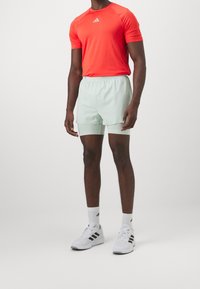 Camiseta deportiva roja de mangas cortas con logo, combinada con pantalones cortos verde menta. Zapatillas blancas con rayas negras y calcetines blancos.