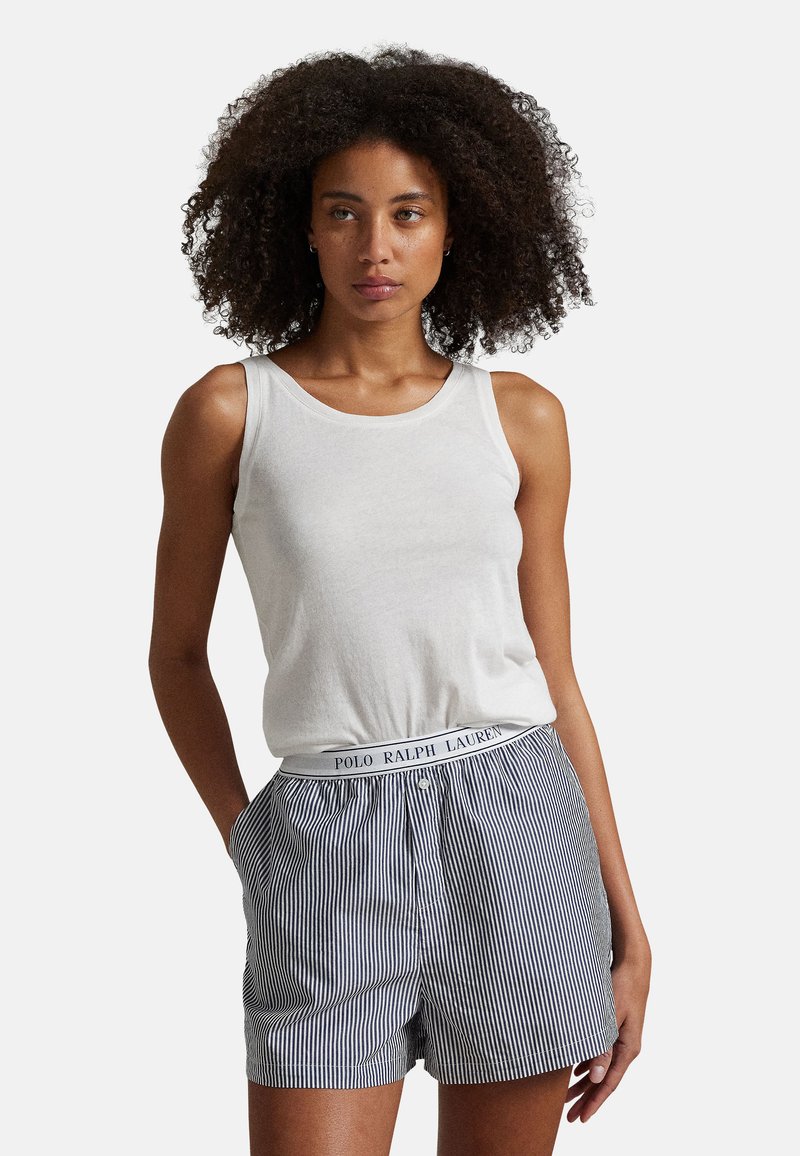 Žena s kudrnatými vlasy, oblečená v bílém tílku a modro-bílých pruhovaných šortkách Polo Ralph Lauren, s rukama v kapsách.