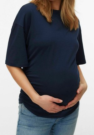 Zwangere vrouw die een marineblauw shirt en spijkerbroek draagt, houdt haar buik voorzichtig met beide handen vast.