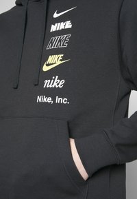 Czarna bluza z kapturem z wieloma logo Nike w różnych stylach i kolorach, nadrukowanymi pionowo na piersi, oraz ręką schowaną w przedniej kieszeni.