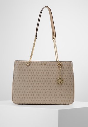 Sac à main beige de créateur avec un motif répétitif du logo DKNY, des anses en chaîne dorée et un charm rond doré avec le logo suspendu à une anse.