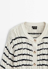 Cardigan blanc en tricot avec des rayures noires horizontales et trois boutons décoratifs dorés sur le devant, étiqueté 100 % coton.