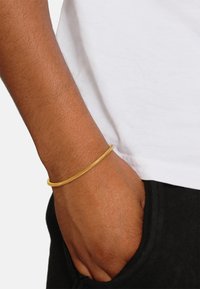 Bracelet en métal de couleur dorée avec un design texturé et courbé, porté au poignet sur un t-shirt blanc et un pantalon noir.