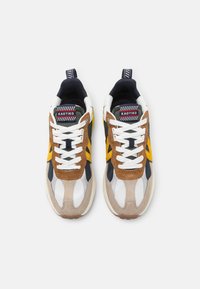 Sportsneaker mit einer Mischung aus beigen, braunen, schwarzen und gelben Materialien, einem strukturierten Design, weißen Schnürsenkeln und einer markierten Zunge.