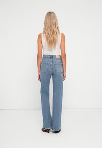 Jeans de cintura alta em azul claro com pernas largas, apresentando dois bolsos traseiros e um patch de couro bege na cintura. Textura de denim suave.