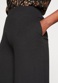 Les pantalons noirs côtelés présentent une taille haute, des poches latérales et une texture lisse. Le tissu est légèrement structuré pour une coupe ajustée.