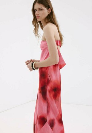 Junge Frau mit langen braunen Haaren, die ein trägerloses pink-rotes Blumenkleid trägt, steht seitlich mit Armbändern am Handgelenk.