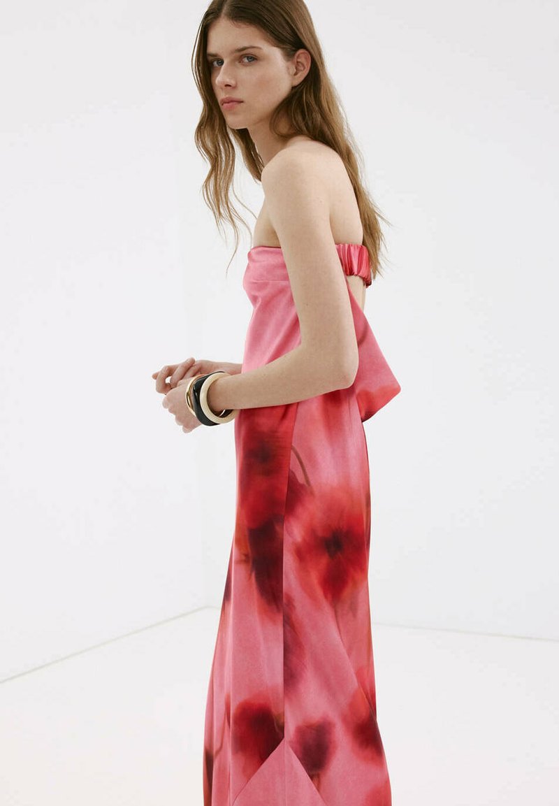 Jeune femme aux longs cheveux bruns portant une robe fleurie rose et rouge sans bretelles, debout de côté avec des bracelets au poignet.