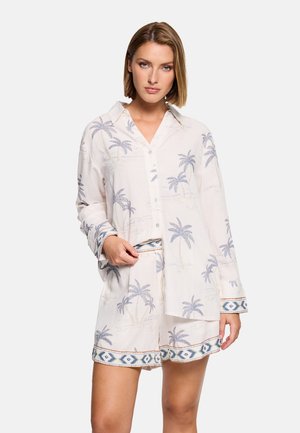 Mujer vestida con una camisa blanca abotonada y pantalones cortos a juego con estampados de palmeras azules y puños y dobladillos con patrones geométricos.