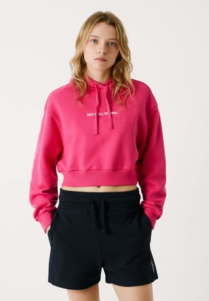 Hoodie rosa a maniche lunghe con cappuccio e cordini, con testo bianco "RED BULL RACING", abbinato a pantaloni corti neri con vita elastica. Tessuto morbido, design casual.