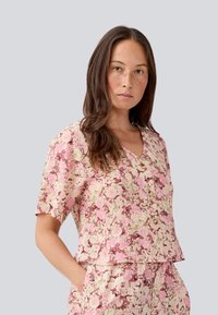 Blusa a maniche corte con collo a V in un modello floreale rosa e crema. Presenta una vestibilità comoda e bottoni decorativi sul davanti. Tessuto morbido al tatto.