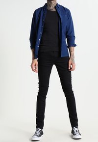 Modrá kostkovaná košile na knoflíky přes černé přiléhavé tričko, doplněné černými úzkými džínami a černými teniskami s bílými detaily.