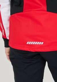 Giacca sportiva rossa e nera con una finitura testurizzata. Presenta accenti riflettenti vicino all'orlo e un pannello a contrasto sulla spalla.