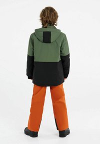 Veste d'hiver verte et noire avec capuche, associée à un pantalon de neige orange. Présente un design bicolore et un tissu durable. Bottes noires visibles.