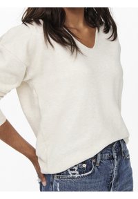 Pull léger beige à col en V avec manches longues, texture douce, coupe décontractée, associé à un short en denim déchiré avec bords effilochés et poches.