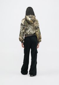 Άτομο με μακριά μαύρα μαλλιά που φοράει ένα cropped hoodie με καμουφλάζ και χαλαρές μαύρες cargo παντελόνες, στέκεται μπροστά από ένα απλό ανοιχτό φόντο.