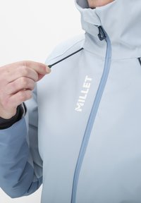 Veste imperméable bleu clair avec col montant, cordon de serrage réglable et logo réfléchissant à l'avant. Texture lisse et design ajusté.