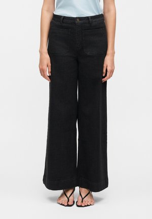 Personne portant un jean noir taille haute à jambes larges avec poches plaquées à l'avant et des sandales noires à lanières, debout devant un fond uni.