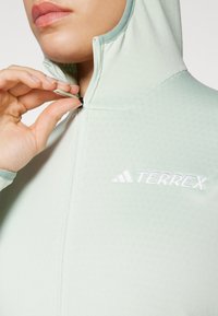 Lys grønn Terrex zip-up genser med teksturert stoff, høy krage og hvit brodert logo på brystet. Hånd som griper glidelåsen.