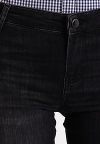 Svarta denimjeans med en slät yta, knappstängning och kontrasterande sömmar. Bärs tillsammans med en rutig blå och vit skjorta.