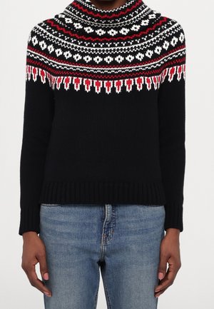 Personne portant un pull en tricot noir avec un motif nordique rouge, blanc et noir sur l'empiècement, associé à un jean bleu clair, debout devant un fond uni.
