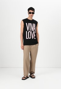 Svart ermeløs t-skjorte med rosa "VIVA LOVE" tekst, beige løstsittende bukser og svarte sandaler. Glatt tekstur og uformelt design.