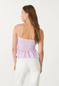 Levandulový peplum top s smockovanou texturou, tenkými ramínky a volánkovým lemem, v kombinaci s bílými džínami. Zadní pohled ukazuje designové detaily.