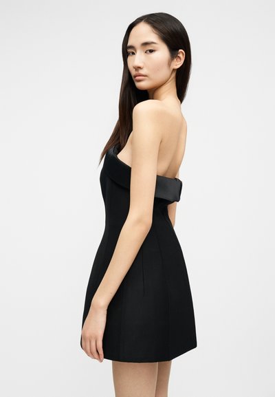 Young woman with long dark hair wearing a strapless black mini dress, standing sideways against a plain light background.