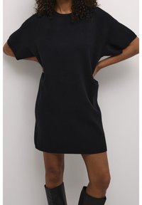 Femme debout, mains sur les hanches, portant une robe en tricot noire à manches courtes et des bottes noires montantes jusqu'aux genoux, sur un fond blanc uni.