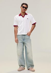 Homme portant des lunettes de soleil noires, un polo en maille blanche avec une bordure bordeaux, un jean large bleu clair et des chaussures beige sans lacets, debout devant un fond uni.