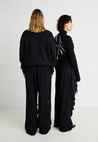 To modeller står ryg mod ryg iført oversized sorte bluser og løse sorte bukser. Den ene bluse har et sølvmønster over ryggen.