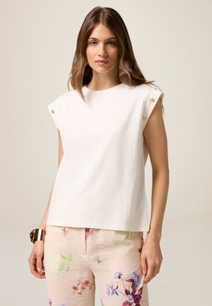 White cotton top with short, structured sleeves and decorative gold buttons on shoulders. Paired with light pink, floral-patterned pants.