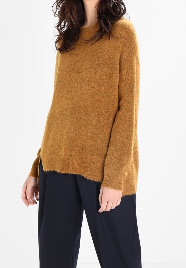 Femme portant un pull ample et duveteux couleur moutarde et un pantalon large foncé, debout devant un fond blanc uni.