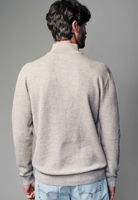 Lysegrå tekstureret sweater med høj krave og ribbede manchetter, præget af et honningkombinationsmønster og en afslappet pasform, parret med lyse denim jeans.
