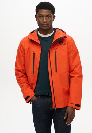 Chaqueta impermeable de color naranja brillante con capucha, cremalleras negras, dos bolsillos en el pecho y puños ajustables. Llevada sobre un suéter de color azul oscuro.