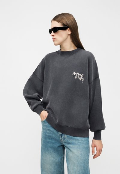Asmuo dėvi per didelį tamsiai pilką džemperį su užrašu „ANINE BING“, šviesiai mėlynas džinsus ir juodus stačiakampius akinius nuo saulės, poziciją palaiko ranką kišenėje.