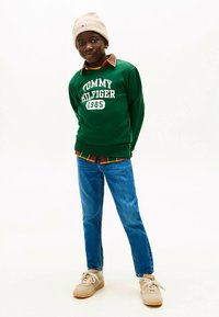 Πράσινο φούτερ με λευκό κείμενο, μπλε τζιν, μπεζ αθλητικά παπούτσια και ελαφρύ μπεζ πλεκτό σκουφάκι. Φορεμένο με πουκάμισο καρό από κάτω.