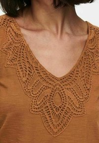 Haut marron à col en V avec des détails en dentelle crochetée symétrique et élaborée autour du décolleté et de la partie supérieure de la poitrine.