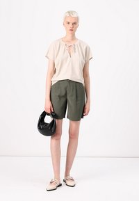 Haut beige à manches courtes avec un col à nouer, shorts olive, sac à main noir et ballerines beiges avec des lanières décoratives.