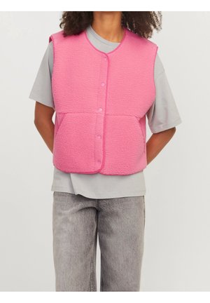 Personne portant un gilet en polaire rose vif par-dessus un t-shirt gris à manches courtes et un jean gris clair, debout les mains dans le dos.
