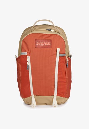 Pomarańczowo-beżowy plecak JanSport z przednią kieszenią na zamek, kieszeniami bocznymi, górnym uchwytem i dwoma pionowymi białymi paskami na przednim panelu.