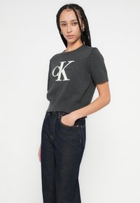 Pulover gri cu mâneci scurte și tiv striat, cu un logo alb proeminent "CK". Asortat cu blugi din denim închis la culoare.