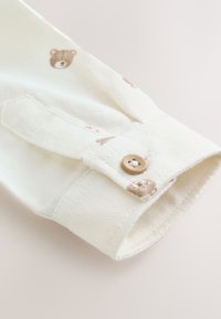 Poignet de chemise en coton crème avec imprimé ours brun. Doté d'une fermeture à bouton et d'un tissu texturé. Détails de couture subtils le long des bords.