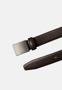 Bruine leren riem met een gladde textuur, voorzien van een rechthoekige metalen gesp met het opschrift "Calvin Klein." Verstelbaar met meerdere gaten.