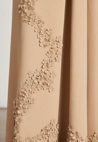 Tissu beige avec des appliqués floraux 3D disposés en un motif tourbillonnant sur un côté, mettant en valeur les détails texturés et le savoir-faire.