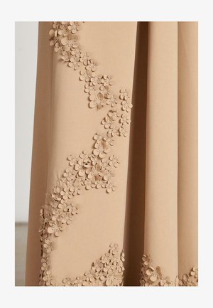 Beige stof met 3D bloemapplicaties die in een wervelend patroon langs één zijde zijn gerangschikt, en die de textuur en het vakmanschap benadrukken.