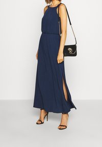 Granatowa maxi sukienka z halterem, plisowany design i bocznymi rozcięciami. Połączona z czarną torebką na ramię z dekoracyjnym okuciem.