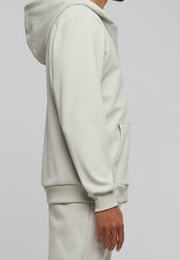 Homme portant un sweat à capuche beige clair et un pantalon de survêtement assorti, vu de profil avec les mains détendues le long du corps sur un fond gris.