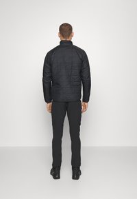 Chaqueta aislante negra con un patrón acolchado, cuello alto y mangas largas, combinada con pantalones y botas negras, mostrada desde atrás.